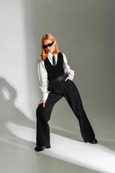 stock image business fashion photography, trendy and red haired asian woman in dark sunglasses, white shirt, black tie, vest and pants posing with hand in pocket on grey shaded background, full length