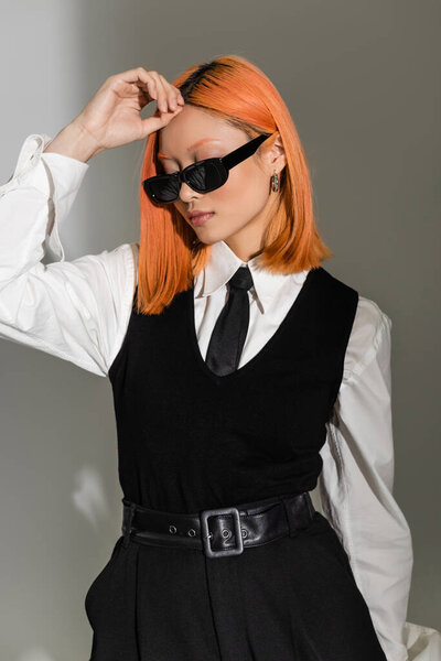 заманчивая азиатская женщина с цветными рыжими волосами в темных солнцезащитных очках, белой рубашке, черном галстуке и жилете, позируя с рукой возле лба на сером затененном фоне, фотографии бизнес-моды