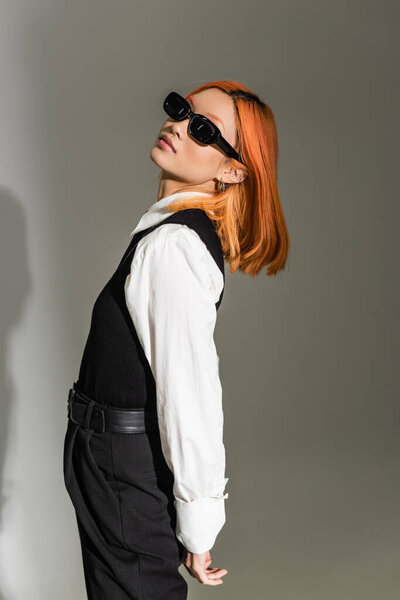 charming and red haired asian woman in dark stylish sunglasses, white shirt and black vest standing and looking at camera on grey shaded background, business casual, generation z lifestyle