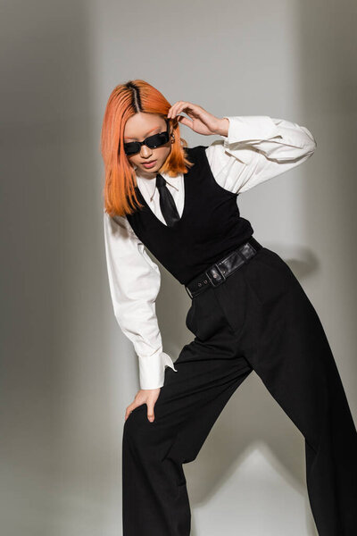 fashion shoot of modern asian woman in dark sunglasses and black and white clothes on grey shaded background, colored hair, white shirt, black tie, vest and pants, stylish pose, business casual