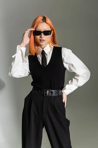 business fashion shoot, young asian woman adjusting dark sunglasses and posing with hand on hip on grey shaded background, white shirt, black pants, tie and vest, modern lifestyle