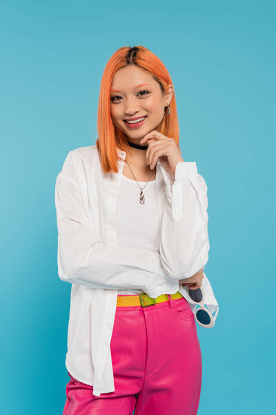 беззаботное лето, положительные эмоции, молодая азиатская женщина с лучезарной улыбкой держа солнцезащитные очки и глядя на камеру на голубом фоне, белая рубашка, розовые брюки, модные аксессуары, поколение z