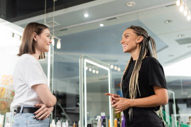 happy beauty worker welcoming female client, cheerful hair stylist with braids talking to smiling woman in salon, customer satisfaction, beauty industry, hair extension, hair colorist, salon job  clipart