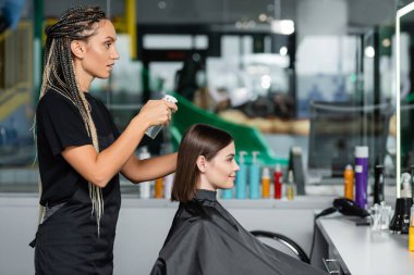 hairstylist spraying hair of female client, hairdresser with braids holding spray bottle near woman with short brunette hair in salon, hair cut, hair treatment, hair make over, hairdo, side view clipart