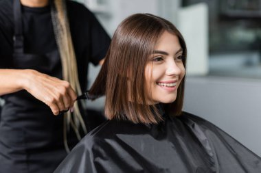 client satisfaction, cheerful woman with short brunette hair sitting in hairdressing cape in beauty salon, getting haircut by professional hairdresser, beauty salon clipart
