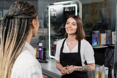 salon services, positivity, tattooed beauty worker in apron welcoming female client with braids in salon, beauty industry, salon job, customer in salon, hairdresser, hair professional  clipart