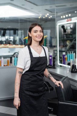 Profesyonel vesikalıklar, önlüklü dövmeli güzellik işçisi güzellik salonundaki kameraya, kuaför koltuğuna, kuaför kuaförüne, saç boyacısına, kuaförlük işine, kuaförlük işine, saç modasına bakıyor. 