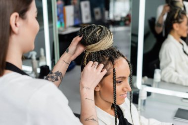 salon experience, tattooed hairdresser doing hair bun to female client with braids, cheerful women, client satisfaction, customer in salon, beauty service, feminine, hair make over, blurred  clipart