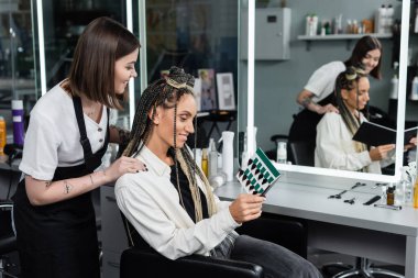 Güzellik salonunda mutlu bir kadın, saç örgülü neşeli bir müşteri dövmeli bir kuaförün yanındaki saç paletine bakıyor müşteri memnuniyeti, saç stili, ayna değişimi, saç tokaları, örgülü saçlar. 