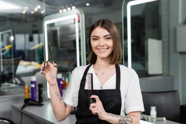 Profesyonel vesikalıklar, pozitiflik, önlüklü dövmeli kuaför elinde saç kesme aletleri, kuaför makası ve tarak, kameraya bakmak, gülümseme, güzellik salonu, kuaför. 