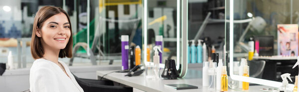 клиент в салоне, татуированная молодая женщина сидит в кресле и смотрит на камеру рядом с инструментами парикмахерской, косметика, наращивание волос, окраска волос, размытый фон, поколение z, баннер 