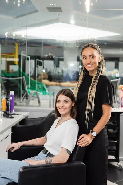 stock image happy hair stylist and female client in beauty salon, beauty worker with braids standing near tattooed woman, looking at camera, hair extension, customer satisfaction, hair care 