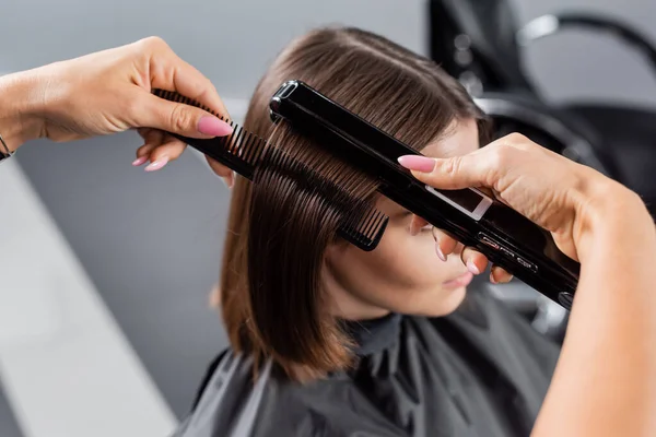 beauty profession, straightening hair, hairdresser with braids brushing and styling short brunette hair of woman in cape, hair smoothing, professional, beauty salon work, hair salon, high angle