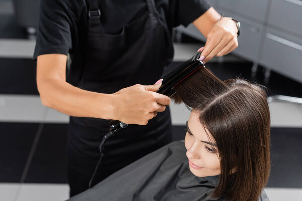 вид сверху профессионального парикмахера с выпрямителем для укладки волос клиентки, косметолога, удовлетворенности клиента, брюнетки с короткими волосами, салон красоты, мода на волосы 