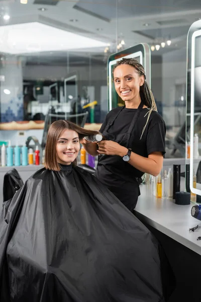 salon services, hairdresser with round brush styling hair of female customer, happy brunette woman with short hair, beauty salon, hair volume, looking at camera, hair professional