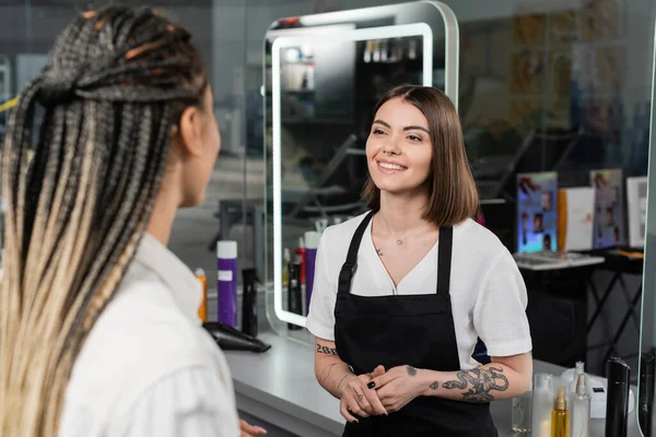 salon services, positivity, tattooed beauty worker in apron welcoming female client with braids in salon, beauty industry, salon job, customer in salon, hairdresser, hair professional