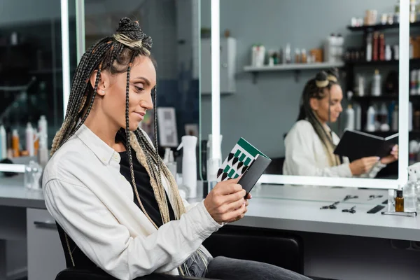 happy woman in beauty salon, joyful client with braids looking at hair palette, customer satisfaction, beauty salon, hairstyle, mirror refection, hair buns, braided hair, hair extension