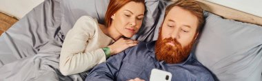 Üst manzara, ağ kurma, rahatlama zamanı, dövmeli çift dinlenme, çocuksuz hafta sonları, karı koca, sakallı adam kızıl saçlı kadının yanında akıllı telefon kullanıyor, rahat yatak odası, ekran saati, afiş. 