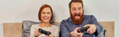 Çocuksuz hafta sonları, kızıl saçlı karı-koca video oyunu oynuyorlar, sakallı adam ve mutlu kadın ellerinde joystickler, heyecanlı, oyun eğlencesi, evli çift, modern yaşam tarzı, afiş. 