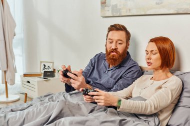 day off without kids, redhead husband and wife playing video game, bearded man and tattooed woman holding joysticks, gaming fun, married couple, modern lifestyle, joy of gaming  clipart