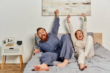 excited people, day off without kids, redhead and happy husband and wife, bearded man and woman with raised hands, parents alone at home, modern lifestyle  clipart