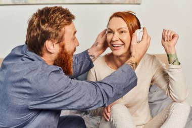 music enjoyment, quality time, day off without kids, redhead husband and wife, bearded man wearing wireless headphones on woman, cheerful parents alone at home, modern lifestyle, relationship  clipart
