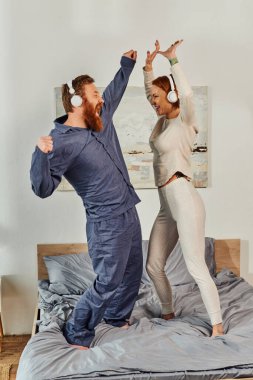 day off without kids, having fun, shared music, redhead husband and wife, happy couple in wireless headphones dancing on bed, bearded man and carefree woman, tattooed music lovers, weekends  clipart