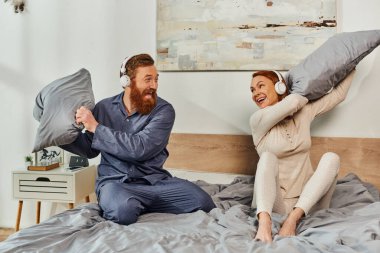 day off without kids, having fun, shared music, redhead husband and wife, happy couple in wireless headphones pillow fight, bearded man and carefree woman, tattooed music lovers, weekends  clipart