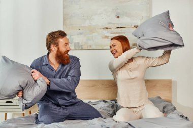 day off without kids, having fun, pillow fight, redhead husband and wife, happy couple in sleepwear enjoying time together, bearded man and carefree woman, tattooed people  clipart