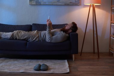 dijital çağ, mobil etkileşim, kızıl saçlı sakallı adam akıllı telefon kullanıyor, kanepede dinleniyor, duvarda resim yapıyor, terlikler halıda, gece, lamba ışığı, boş zaman, rahat yaşam, yan görüş 
