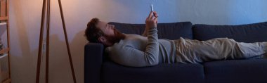 Mobil etkileşim, kızıl saçlı sakallı adam akıllı telefon kullanıyor, kanepede dinleniyor, duvarda resim yapıyor, terlikler halıda, gece lambanın ışığı, boş zaman, rahat yaşam, dijital çağ, afiş. 