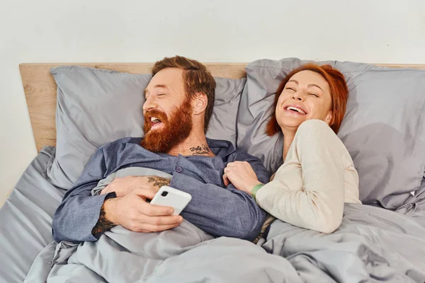 笑いとリラックスタイム子供のいないタトゥーカップル休日夫と妻髭の男と赤毛の女性スマートフォンユーザー居心地の良いベッドルーム屈託のない画面時間 — ストック写真