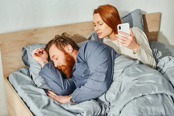 stock image quiet house, parents alone at home, redhead wife looking at husband, bearded man sleeping near woman using smartphone, networking, day off, wake up, tattooed, couple without kids 