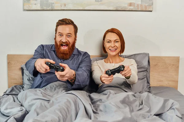stock image weekends without kids, redhead husband and wife playing video game, bearded man and happy woman holding joysticks, excited, gaming fun, married couple, modern lifestyle 