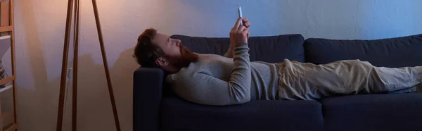 スマートフォンを使って赤い髪の髭を生やした男がソファに座り壁に絵を描くカーペットにスリッパ夜ランプからの光余暇時間居心地の良い生活デジタル時代バナー — ストック写真