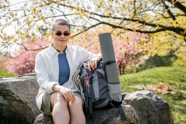 Positive short haired and young female hiker in casual clothes and sunglasses sitting near backpack with fitness mat on stone with blurred nature at background, confident female explorer clipart