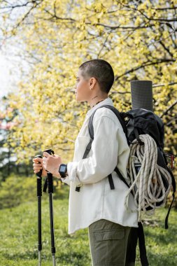 Side view of young short haired and tattooed female hiker with backpack and climbing rope holding trekking poles and walking on blurred nature, independent traveler embarking on solo journey clipart
