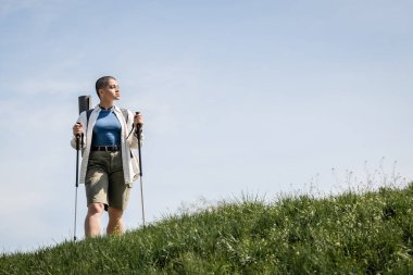 Young short haired woman hiker in casual clothes with backpack holding trekking poles and standing on grassy hill and sky at background, explorer woman discovering hidden trails clipart