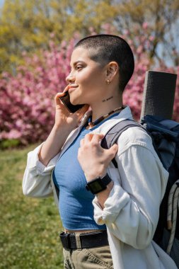 Side view of young short haired and tattooed woman hiker with backpack talking on smartphone while standing with nature at background, curious hiker exploring new landscapes clipart