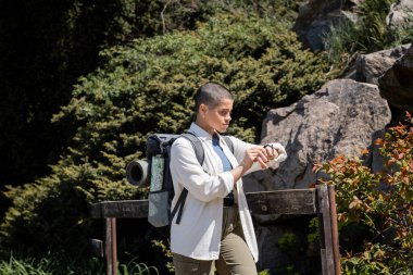 Young short haired female traveler in casual clothes with backpack looking at wristwatch and standing near fence with nature at background, woman trekking across vast landscapes clipart