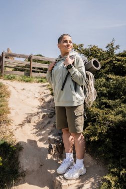 Smiling and short haired female hiker with backpack, fitness mat and climbing rope standing and looking away on hill with sky at background, hiker finding inspiration in nature clipart