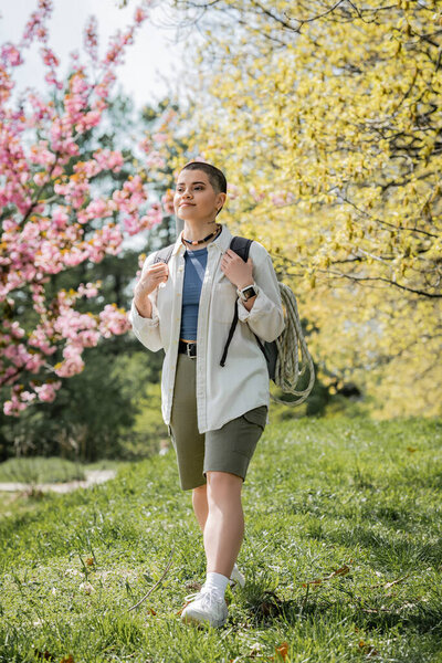 Smiling short haired and tattooed female hiker in casual clothes holding backpack while walking on grass with landscape at background, independent traveler embarking on solo journey