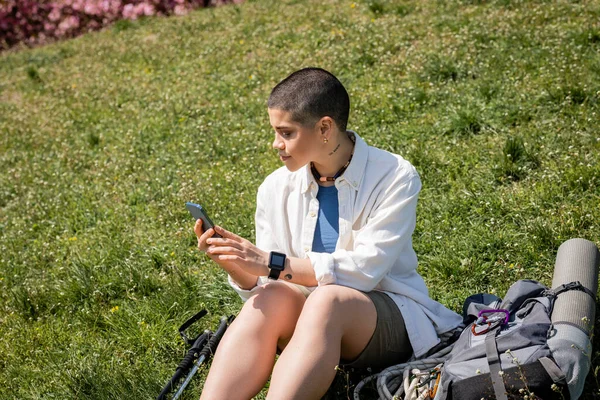 stock image Young short haired and tattooed hiker in casual clothes using smartphone while sitting and relaxing near trekking poles and backpack on grassy hill, explorer woman discovering hidden trails