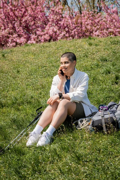 stock image Cheerful young short haired traveler talking on smartphone while sitting near trekking poles and backpack on grassy hill with nature at background, curious hiker exploring new landscapes