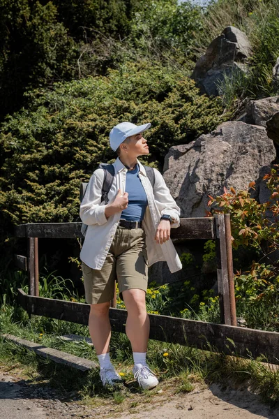 屋外のフェンスの近くに立っている間 バックパックとカジュアルな服や野球帽で若い短髪の女性旅行者のサイドビュー タトゥーの翻訳 — ストック写真