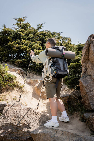 Young short haired female traveler with backpack walking with trekking poles on hill with stones and grass at background, tranquil hiker finding inner peace on trail, summer
