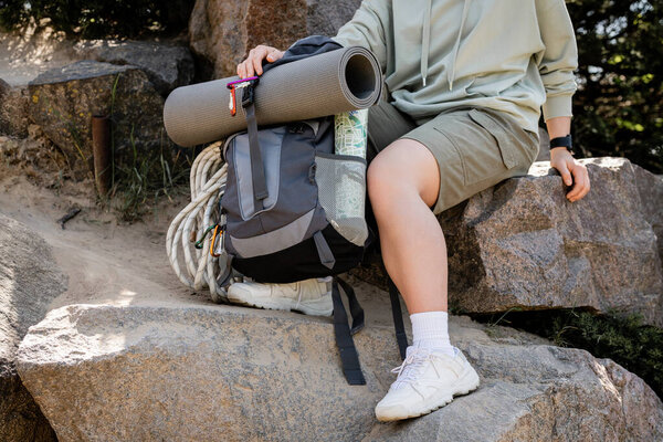 Cropped view of young woman in casual clothes sitting near backpack with map and climbing rope on stones and hill at nature, tranquil hiker finding inner peace on trail, summer