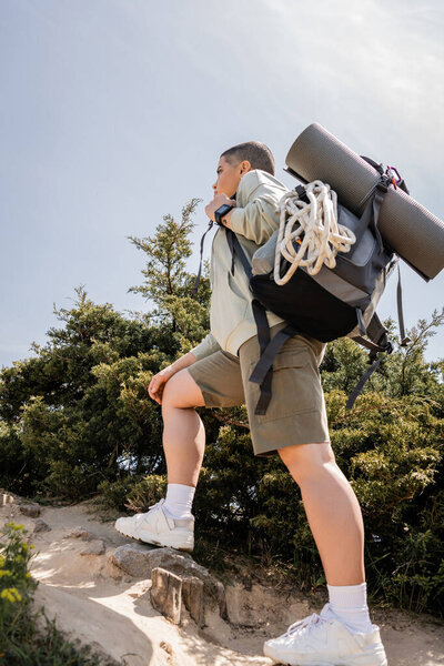 Low angle view of young short haired traveler with backpack and climbing rope walking on hill with stones and nature at background, hiker finding inspiration in nature, summer