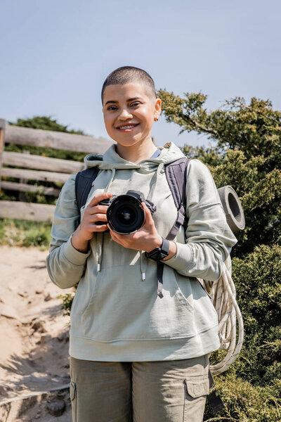 Cheerful young short haired female tourist with backpack and travel supplies holding digital camera and looking at camera with nature at background, hiker finding inspiration in nature