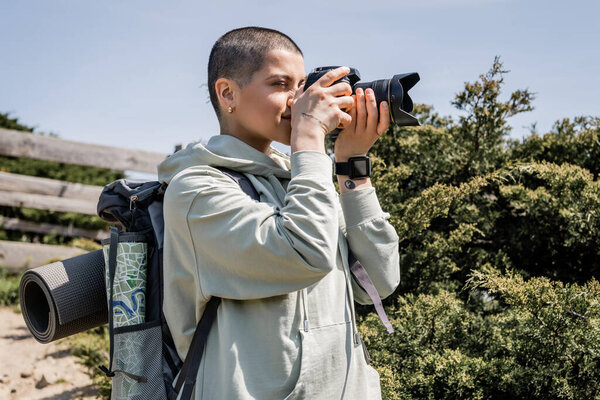 Young tattooed and short haired female tourist with backpack and map taking photo on digital camera while standing with nature at background, travel photographer, summer, Translation of tattoo: love